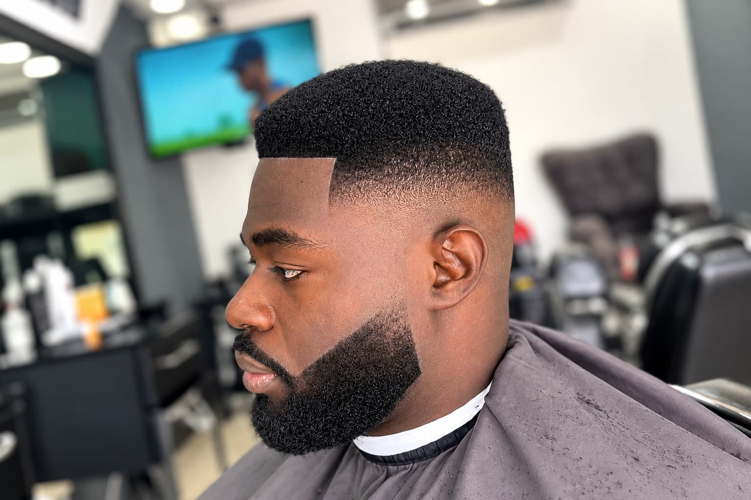 Taper fade with a carved side parting and a sharply lined beard, side profile in the chair